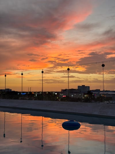 This image captures a sunset over the city of Mérida, Mexico, viewed from what appears to be a rooftop pool or balcony. The sky is ablaze with vibrant orange and pink clouds, casting a warm glow over the scene. In the foreground, a swimming pool reflects the colorful sky and the silhouettes of a few thin, vertical poles, possibly part of a fence or barrier, and a blue floating object. Beyond the pool, a low-rise cityscape is visible, characterized by buildings and illuminated signs suggesting commercial activity. The overall impression is one of urban tranquility at dusk, with the natural beauty of the sunset complementing the man-made environment.