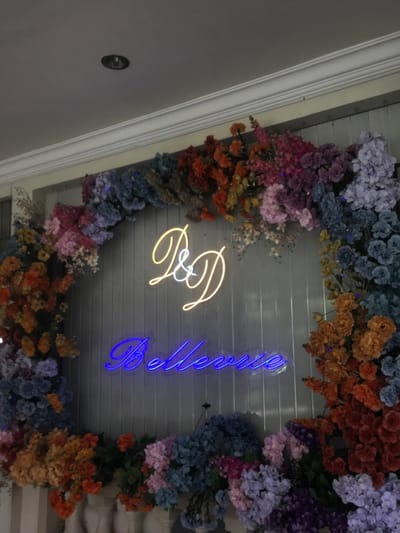 An indoor scene in Kota Batu, Indonesia, depicts a decorative wall display. The central feature is a light-colored, vertically paneled wall adorned with an arch-shaped arrangement of artificial flowers. The flowers exhibit a variety of colors including shades of pink, purple, blue, orange, red, and yellow, forming a vibrant frame. Mounted on the wall within this floral arch are two illuminated neon signs. The upper sign displays the cursive initials "P & D" in yellow-orange neon. Below it, the word "Bellevue" is spelled out in cursive blue neon. Above the display, a white ceiling with decorative crown molding is visible, along with a single recessed light fixture. Partially visible at the bottom of the image are white, decorative balusters, suggesting a railing or structural base. No individuals are present in the image.