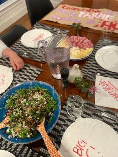 The image shows a table setting in Rye, New York, USA, located at the coordinates 40.952 latitude and -73.708 longitude. The table is covered in a wooden surface with a black and white checkered pattern. In the center of the table are two place settings. One place setting includes a blue plate with salad. The salad appears to have greens, grains, dried fruit, and nuts. Next to the salad is an orange and yellow serving spoon. On the other side of the table is a partially eaten plate with a white napkin. The plate contains several pieces of salami and cheese. There is also a partially filled pitcher of water. Next to the pitcher is a small ceramic rooster. On the opposite side of the table, there are two white plates with napkins. One napkin has the word "Concord" written in red and the other has the word "Big" written in red. There is also a piece of paper on the table that appears to have the word "FARMS" written on it. An arm and hand are visible in the lower left corner of the image. A few glass containers appear to be on the table as well. The setting is a formal dinner.