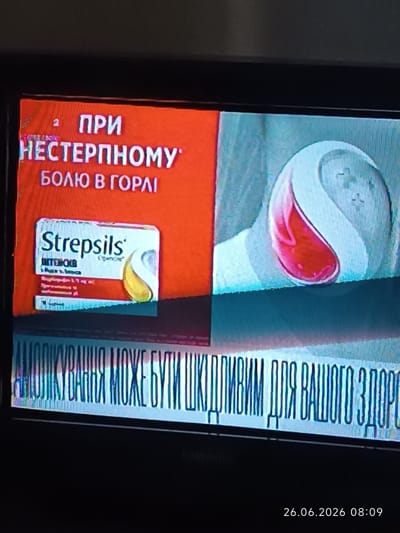 This is a screenshot from a television screen, likely showing a commercial or public service announcement. The left side features an orange background with text in white. The text reads: "ПРИ НЕСТЕРПНОМУ БОЛЮ В ГОРЛІ" which translates to "FOR INTENSE SORE THROAT". Below this, a box of "Strepsils" lozenges is prominently displayed. The right side of the screen shows a stylized graphic of a throat lozenge, appearing as a large, pale blue shape with a pink liquid drop at its center.

A horizontal banner of text runs across the bottom of the screen, largely obscured by digital artifacts and low resolution, but appears to be in a Cyrillic alphabet and likely conveys a warning or informational message about health. Below this, the date and time "26.06.2026 08:09" are visible, indicating the time of recording or broadcast. The overall impression is of a health-related advertisement.
