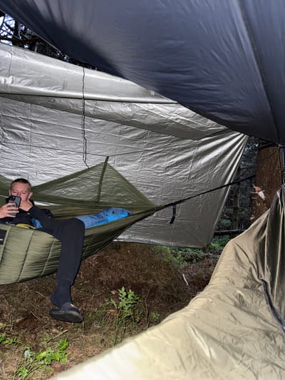A young man wearing a black jacket and black pants is lying in a green hammock. The hammock is suspended between two trees in Macclesfield Forest in the United Kingdom. The man is looking at a phone that he is holding in his right hand. The hammock is covered by a large tarp held up by a rope and a tree. The tarp is a light grey, and there is another tree in the background. The man is lying in the hammock with his legs bent. He is wearing black socks and dark shoes. There is green foliage on the forest floor. The forest is dark and appears to be wooded.