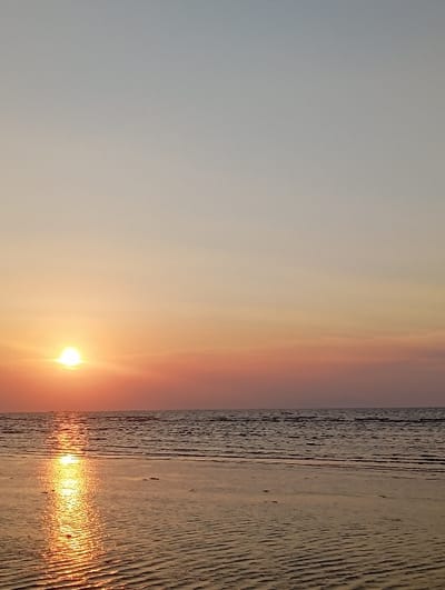 The image depicts a sunset over a coastal area in the city of Tritunggal, Indonesia. The sky, occupying the upper two-thirds of the frame, transitions from a pale blue-grey at the zenith to vibrant orange and pink hues near the horizon. The sun, a bright, circular, golden-orange orb, is positioned on the left side, close to the horizon. Its intense light creates a luminous, shimmering reflection path that extends across the surface of the water and onto the wet sand in the foreground. The middle ground features a body of water, likely an ocean or sea, exhibiting subtle ripples. The immediate foreground is composed of exposed wet sand, clearly showing distinct, parallel ripple patterns. No individuals, animals, or man-made structures are present in the scene, and no specific actions or interactions are observed.