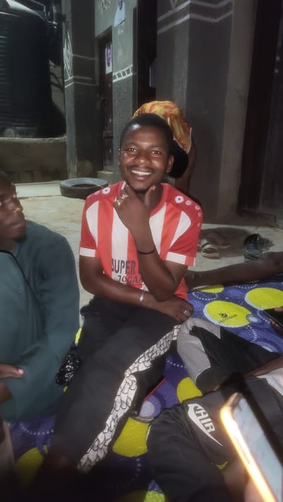 The image depicts a group of people in an indoor setting, presumably in Jogana, Nigeria. A man in a red and white striped shirt is smiling at the camera and making a gesture with his hand. He is wearing a ring on his finger and a bracelet on his wrist. To his right are the legs of people, sitting on a colorful mat. Some are wearing black pants with the word "AIR" on them. There is a phone with a yellow glow visible in the upper right. Other people are sitting, and a bit of another person is visible in the bottom of the image. In the background there are indoor objects.