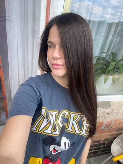 A young woman with long, dark hair is pictured from the chest up, looking to her left. She is wearing a dark blue t-shirt with a Mickey Mouse graphic. She has a nose ring. She is standing on a balcony or patio, with a brick wall visible behind her and a window with curtains and a plant in the background. The scene appears to be outdoors. The location is the city of Romny, Ukraine.