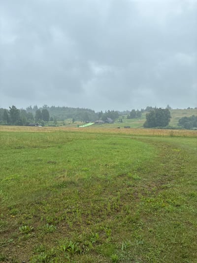 The image depicts a cloudy, overcast day in the city of Sabile, Latvia. In the foreground, there is a wide-open field of green grass with some patches of yellow wildflowers. The grass appears somewhat uneven and mowed. In the middle ground, there is a dirt path or road leading towards a cluster of buildings and trees. One building appears to be a dark-colored house, with a white car parked in front of it. Further to the right, there is a bright green structure, possibly a ramp or slide, set against a backdrop of trees and rolling hills. The background consists of a dense forest of green trees covering the hills, under a heavy, gray sky. The overall impression is of a rural or semi-rural landscape on a day with poor weather.