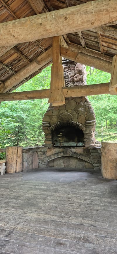 This is a medium shot, vertical, interior view of a rustic outdoor pavilion with a stone barbecue. The pavilion has a roof constructed of wooden beams and planks, with exposed rafters. A large stone chimney and barbecue grill dominate the center of the image. The grill appears to be made of metal. To the left and right of the barbecue are large, rough-hewn wooden logs. The floor of the pavilion is made of weathered wooden planks. In the background, a dense forest of green trees is visible. The scene appears to be located outdoors in a natural setting, possibly a park or campsite, and there are no people present. The image does not contain information that would indicate its location in Knevichi, Russia.
