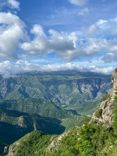 From a high vantage point, the photograph captures a mountainous landscape with lush green vegetation covering the terrain. A rocky cliff edge is visible in the foreground. The sky is partially cloudy, with patches of blue sky visible among the white and gray clouds. This image likely comes from the Aranđelovo area of Bosnia and Herzegovina given the vegetation and terrain.