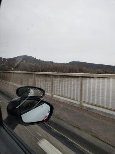 The image depicts a scene observed from inside a moving vehicle on an overcast day, looking across a body of water towards Rochemaure, France. In the lower left, a black car's side mirror is prominently visible, featuring an attached smaller, oval-shaped convex blind spot mirror. Raindrops and streaks are present on the vehicle's window. Below the mirror, a wet, dark asphalt road surface with faint white lane markings shows motion blur. A light-colored concrete bridge barrier with a metal railing composed of vertical bars extends horizontally across the mid-ground. Beyond this railing, a body of water, likely the Rhône River, reflects the vertical bars of the railing on its choppy surface. The far bank of the river is lined with a dense growth of bare deciduous trees. In the background, dark, undulating hills rise, upon which several structures, consistent with buildings or a castle, are visible, forming part of the city of Rochemaure.