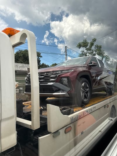 A maroon Hyundai Tucson is being transported on a white flatbed tow truck. The car is positioned facing forward on the truck's bed. The tow truck has an orange beacon light on its top front left side. The truck's side has a printed hotline number: "Hotline: 0338 330 777". The background shows a residential area with houses, trees, and power lines under a cloudy sky. The scene appears to be in a city, possibly Tu Póh, Vietnam, based on the context provided. The image is taken from a slightly elevated angle, looking towards the car and tow truck.
