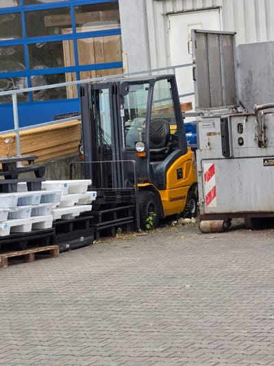A yellow Jungheinrich forklift is parked next to a large grey industrial container or vehicle. The forklift has a black seat and a metal cage surrounding the driver's area. Its front wheels are visible, and the tires show signs of wear. Behind the forklift, stacked on pallets, are several white and light grey objects that appear to be plumbing fixtures or bathtubs. These are partially obscured by black metal racks and wooden pallets.
The scene appears to be an outdoor industrial or storage area. The ground is paved with a textured grey material, possibly a permeable paver. Green foliage and some dry leaves are visible around the base of the forklift and container, suggesting a natural element in an otherwise man-made environment.
To the left of the forklift, a section of a building with blue-framed windows is visible. Some of the windows reflect the surroundings, showing what might be parked vehicles or other structures. Wooden planks are stacked next to the building.
A sign with red and white diagonal stripes is affixed to the grey container, along with other labels. Text is visible on the forklift, reading "JUNGHEINRICH." Another label on the container includes German text, likely related to safety or identification.
The lighting suggests it is daytime, and the weather appears to be overcast, given the diffused light and lack of strong shadows. The overall impression is a functional, industrial setting.