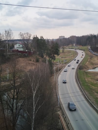 This image captures an elevated view of a winding road traversing a semi-urban landscape in Khotkovo, Russia, on an overcast day, likely in early spring or late autumn given the predominantly bare trees.
In the foreground, dark, bare tree branches frame the bottom left, and prominent power lines or cables stretch horizontally across the top portion of the frame. A body of dark water, possibly a river or pond, is partially visible among the trees in the lower left.
The midground is dominated by an asphalt two-lane road that curves through the scene from the lower left towards the upper right. Several cars, mostly dark-colored sedans and SUVs, are visible on the road, indicating active traffic. To the left of the road, on a small hill, stands a light green house with a distinct red roof, surrounded by a red fence and bare deciduous trees. Further along the road, green grassy embankments line its sides, and a grey pedestrian bridge arches over a lower area in the upper right. Utility poles are scattered throughout the landscape.
In the background, distant hills covered with dense, bare forest give way to faint outlines of multi-story buildings, suggesting a town or city center beyond the immediate natural area. The sky is completely covered with light grey clouds, creating a diffused light with no harsh shadows. No specific activity beyond regular traffic is depicted, and no discernible text is visible on any objects or signs.