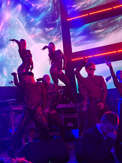 The image shows a group of performers on a stage, illuminated by bright stage lights. The performers are wearing shiny, dark costumes. The background features a large screen displaying an abstract, colorful image with a galaxy-like pattern. Red and blue lights are prominent in the scene, creating a vibrant and energetic atmosphere. In the foreground, a person in a suit and glasses is seated, appearing to be watching the performance. The setting is likely an indoor venue. There is no specific indication of the location being in the city of Tsilkani, Georgia.