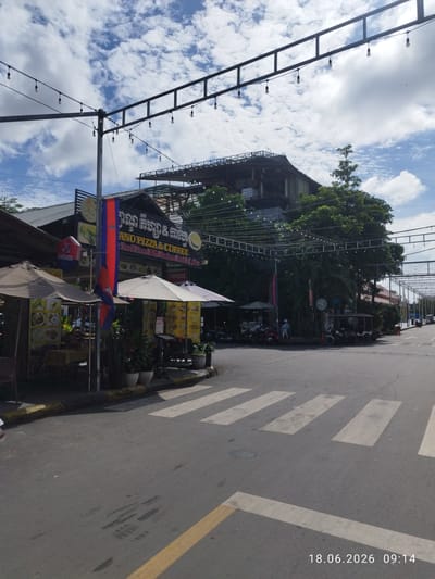 The image depicts a street scene in Kampot, Cambodia, on a partly cloudy day. In the foreground, a crosswalk is visible on an asphalt road. To the left, a restaurant with outdoor seating is set up under umbrellas, featuring menus displayed on the tables and walls. A Cambodian flag is prominently displayed on a pole. Behind the restaurant, the facade of a building with signage for "ANO PIZZA & COFFEE" and "Mexican Food & Coffee" is visible. Overhead, strings of lights are suspended across the street. In the background, trees and other buildings can be seen, and a few tuk-tuks are parked on the side of the road. The timestamp "18.06.2026 09:14" is present in the lower right corner of the image.