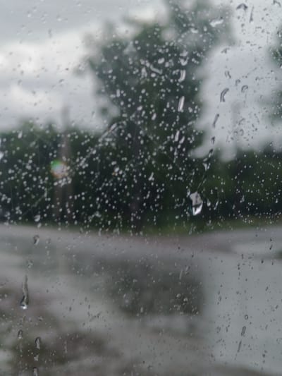 This media file captures a view through a wet window, likely during rainy weather. The foreground is dominated by numerous raindrops clinging to the glass, distorting the view of the scene beyond. Some drops are small and circular, while others are elongated, suggesting they are running down the pane.

The background is blurred due to the water on the window and the shallow depth of field. It appears to be an outdoor scene with trees and foliage in a muted green color. The sky is overcast and gray, typical of rainy conditions. There is a hint of a road or pathway in the middle ground, also appearing wet and reflecting the dull light.

The overall impression is one of a gloomy, overcast day with rain. The location context suggests this scene is from Ақжар, Kazakhstan. There is no visible text within the image. The scene evokes a sense of melancholy or tranquility often associated with rain. The specific time of day is not clearly discernible but appears to be daytime.