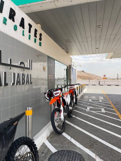 A line of orange and white dirt bikes is parked outside a building with “EMIRATES” and “AL JABAL” inscribed on its facade. The building appears to be near a mountain and a parking zone marked with white stripes. A manhole cover is visible on the right side of the image. The sky is overcast.