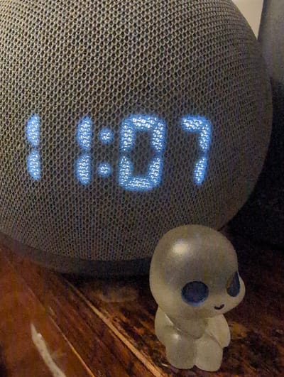 The image features a round, mesh-covered smart speaker displaying the time "10:37" in blue digital numerals. A small, translucent white duck figure with blue eye spots is positioned to the right of the speaker. The background includes what appears to be a dark wooden surface.