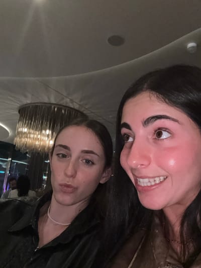 The image displays two young adult females captured in a close-up indoor setting within an upscale establishment in the city of Rum Cay, Bahamas. The woman positioned on the right has dark hair and brown eyes, looking slightly upwards and to the right with a wide, open-mouthed smile, revealing her teeth. Her complexion shows a reddish flush, and she is wearing a light brown top with a faint chain necklace. The woman on the left has dark hair and blue-grey eyes, maintaining direct eye contact with the camera, exhibiting a neutral expression with slightly pursed lips. She is dressed in a dark, collared jacket and a thin, white beaded choker necklace. The background features a large, modern chandelier with multiple vertical, translucent elements, illuminating a curved, light-colored ceiling. A recessed ceiling speaker is visible in the upper right. A blurred figure of another individual is partially discernible in the left background, facing away from the camera. The scene is illuminated by dim, artificial ambient lighting.