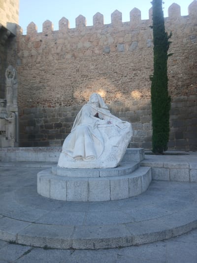 A full-shot, eye-level, outdoor, daytime photograph shows a white stone sculpture of a cloaked figure seated on a rough-hewn rock base, which is itself situated on tiered, grey stone steps. The sculpture depicts a figure with a serene expression, wearing a hooded garment that drapes over their body, with hands resting on their lap. A small bunch of red flowers is visible in the figure's hands.
Behind the sculpture, a tall, dark green cypress tree stands next to a thick, rough-hewn stone wall that forms the background. This wall has battlements along its top edge. To the left of the sculpture, another stone structure, possibly a building or monument, is partially visible, with faint carved details on its surface. The ground in the foreground is paved with grey flagstones, some of which are curved. The overall scene suggests a historical or religious setting, likely in the city of Ávila, Spain, given the architectural style.