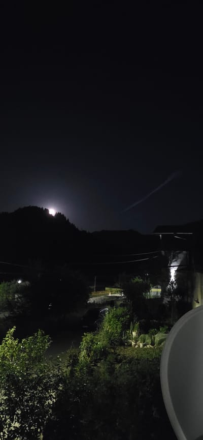 A nighttime landscape view, likely taken from an elevated position. A dark sky occupies the left portion of the frame, with a contrail visible in the upper portion. In the middle is a dark, silhouetted mountain range. The moon is visible shining over the mountain range. The right part of the image shows a residential area with vegetation, trees, and a road. A white, unidentified object is visible in the upper right corner. The location is potentially Rudozem, Bulgaria, based on the user's request.