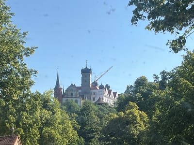 The image depicts a large, multi-towered building, likely a castle or monastery, situated atop a wooded hill. The architecture features spires, turrets, and various rooflines, with a prominent clock tower and a construction crane visible against the sky. Lush green trees surround the building, obscuring some of its lower sections. The sky is clear and blue, indicating a bright, sunny day. The presence of the crane suggests ongoing construction or renovation work. The scene is a picturesque view of a historical structure nestled within a natural landscape, characteristic of a European setting like Scheinfeld, Germany. The overall mood is peaceful and serene, despite the visible construction activity. No text is discernible in the image.