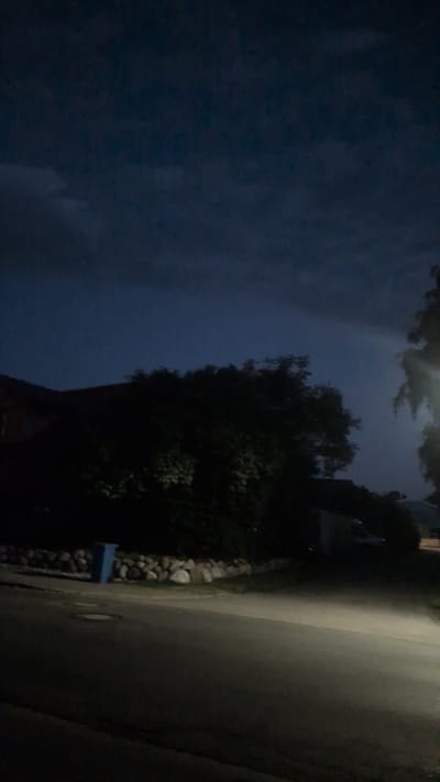 The image depicts a nighttime street scene in the city of Hetlingen, Germany. The sky is dark and overcast, with some lighter clouds visible. In the midground, a cluster of trees is silhouetted against the dark sky. A low stone wall runs along the edge of a property, and a blue trash bin is visible behind it. A street stretches across the foreground, with a vehicle's headlights illuminating the road ahead. The overall impression is one of a quiet, possibly residential, area at night.