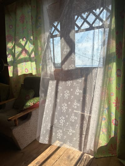The image depicts an interior scene of a room, possibly a sunroom or porch, with light filtering through a window and casting shadows. The room appears to be in a rustic setting, possibly a dacha in Berkuty, Russia, given the wooden floorboards and rustic window frame.
In the foreground, on the left, is a portion of an armchair upholstered in a patterned fabric. A few decorative pillows are placed on the chair, one with a floral pattern in shades of green and pink. The floor is made of wooden planks.
Dominating the center and right of the image are several curtains. On the far left, a green curtain with a floral pattern featuring pink, blue, and yellow flowers is visible. In the center, a sheer white lace curtain with a delicate floral pattern hangs, partially obscuring the view through the window. On the far right, another green curtain with a similar floral pattern is seen.
The window, partially covered by the sheer curtain, has a rustic wooden frame with a diamond-shaped lattice pattern. Through the window, a bright sky with scattered clouds is visible, suggesting daylight. Sunlight streams through the window and the curtains, creating strong diagonal shadows across the floor and on the curtains themselves.
There are no people visible in the image. The overall impression is one of a peaceful, sunlit, and somewhat cozy domestic interior.