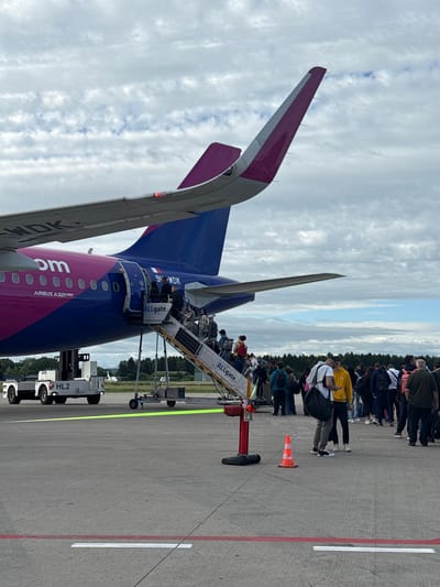 A wide shot captures a Wizz Air Airbus A321neo aircraft on the tarmac at an airport, likely in Künersberg, Germany, given the context provided. Passengers are boarding the plane via a mobile staircase, with a significant number of people visible both on the stairs and gathered on the ground. The aircraft's distinctive purple and blue livery is prominent. A tow tractor labeled "HL2" is parked to the left of the plane. The sky is overcast with scattered clouds. The scene depicts a typical airport boarding process.
