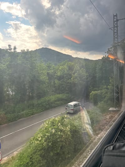 The image, captured from a moving vehicle, overlooks a two-lane asphalt road winding through a densely wooded, hilly landscape. A silver station wagon is observed traveling on the road. The surrounding environment is characterized by lush green trees and vegetation, with a mountain range forming the backdrop under a cloudy sky. The scene suggests a rural or semi-rural setting, possibly in the vicinity of Zlidol, Bulgaria, given the mention of the location. A tall, latticed metal pole, likely part of an electrical transmission tower, is visible on the right side of the frame. Reflections on the window indicate the perspective is from inside a train or a similar mode of transport.