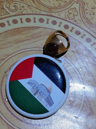 A gold ring with a dark red stone sits next to a circular plastic keychain. The keychain displays an image of the Dome of the Rock over the Palestinian flag. The ring and keychain rest on a surface with a patterned design. The colors appear slightly faded, and the composition includes a close-up perspective.