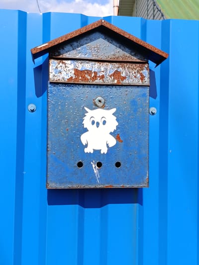 The image shows a weathered blue metal mailbox attached to a bright blue corrugated metal fence. The mailbox has a rustic, house-shaped roof made of rusted metal. A white sticker of a cartoon owl is affixed to the front of the mailbox. Below the owl sticker, there are three small holes in the mailbox. The mailbox is mounted to the fence with two visible bolts. The scene is outdoors, with a hint of sky and greenery visible at the top of the frame. The lighting suggests it is daytime. The location is specified as Смілавіцкі сельскі Савет, Belarus.