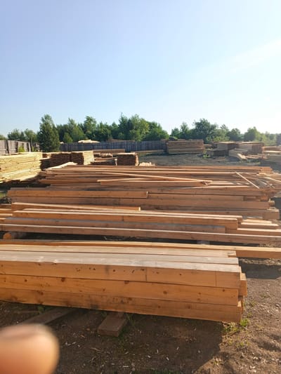 The image shows a lumber yard under a clear blue sky. Stacks of raw lumber, primarily pine, are arranged in neat piles. In the foreground, large, square-cut timber beams are stacked horizontally. Behind these, thinner planks are arranged in several large, organized stacks. The lumber yard is situated outdoors, with a wooden fence visible in the background and a line of trees beyond the fence. The ground is composed of dirt and some scattered debris. The scene suggests a processing or storage area for building materials. The location is identified as Strelka, Russia. There are no people visible in the image.