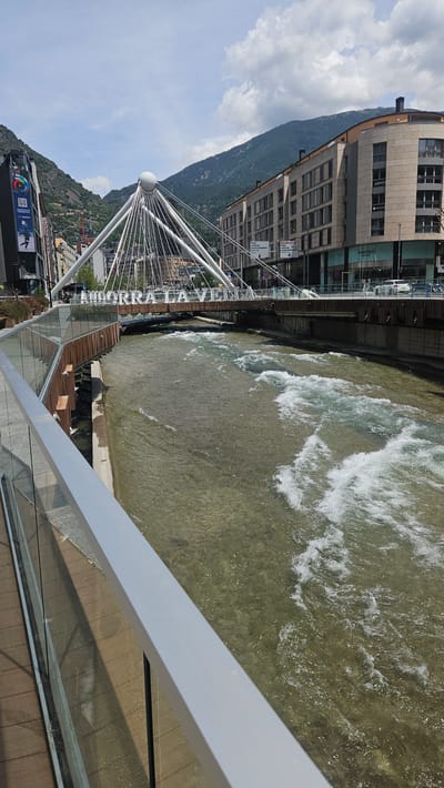This image captures a vibrant urban scene in Andorra la Vella, Andorra, on a clear, sunny day with some scattered clouds. The primary subject is the Valira river, flowing from left to right, characterized by visible rapids and a clear, shallow appearance with a light brown, rocky bed.
In the foreground, a modern pedestrian walkway is visible, featuring a grey metal railing and a transparent glass barrier extending diagonally from the lower left to the mid-right. The walkway surface appears to be composed of light brown wooden planks.
Spanning the river in the midground is the distinctive Pont de la Rotonda (also known as Pont de París), a cable-stayed bridge. This bridge is architecturally notable for its central white spherical element from which numerous white cables radiate downwards to support the bridge deck. Large white capital letters on the bridge's framework spell out "ANDORRA LA VELLA." The riverbanks feature a mix of modern retaining walls, some appearing to be wooden or metallic on the left, and concrete or stone on the right.
In the background, on either side of the river and bridge, are multi-story urban buildings. To the left, a tall, dark building features a large vertical digital display with colorful graphics. Behind this, a steep, tree-covered mountain rises. To the right, a large, modern, curved building with numerous windows on multiple floors is prominent, with "MANGO OUTLET" signage visible on its ground level. A street runs in front of this building, with several white cars parked or in motion. Beyond the buildings, another large, forested mountain range dominates the horizon under a partially cloudy blue sky. A few indistinct figures are visible on the bridge and streets.