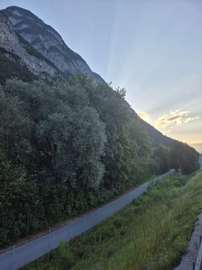The image captures a mountainous landscape adjacent to a paved path, possibly a bike or walking trail, in Kematen in Tirol, Austria. A steep, rocky mountain face dominates the left side of the frame, partially covered by dense green trees. The path runs diagonally from the bottom left towards the upper right, bordered by a metal fence on the left and overgrown grass and vegetation on the right. The sky above is a soft blue with faint rays of sunlight breaking through from behind the mountain, suggesting it is either early morning or late afternoon. There are no people or animals visible in the scene. The overall impression is one of a natural, somewhat remote area within or near a town.