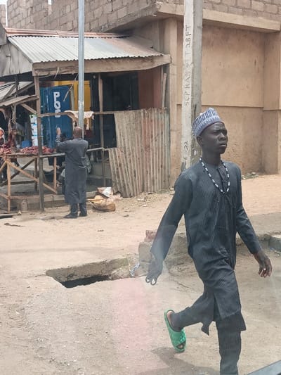 This scene is located on a dusty street in Hotoro North, Nigeria, under daylight conditions. In the foreground, a dark-complexioned male, estimated to be in his 20s or 30s, walks from right to left. He wears a dark grey striped long-sleeved tunic over matching trousers, a blue and white patterned kufi cap, and a black beaded necklace. He has a single green slide sandal on his raised left foot and holds a small, light-colored object in his left hand. His gaze is directed forward and slightly upward.
In the midground, to the left, a bald male in dark grey traditional attire stands with his back to the viewer, facing a prominent blue cabinet or refrigerator marked with "PO". His arms are raised, suggesting interaction with the cabinet. Adjacent to him on the left, an informal market stall displays cuts of raw meat on a wooden table structure. Further left, a structure with a corrugated metal roof is visible.
A utility pole, marked with "SCP RAN LT 28FT" and "6-22", stands vertically in the midground, positioned between the walking man and the shops. The immediate ground is uneven, dusty dirt, featuring an exposed concrete-lined ditch or drain opening in the foreground. In the background, buildings constructed from unplastered concrete blocks are visible, along with corrugated metal siding on some structures.