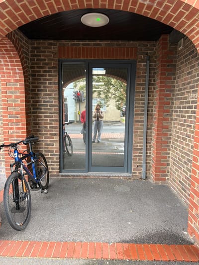 The image depicts the entrance to a building in Mole Valley, United Kingdom. The entrance is framed by a brick archway leading to a gray-framed glass door, which reflects a person taking a picture and a street scene with a traffic light. A blue and black bicycle is parked haphazardly in front of the door. The ground is paved with dark gray asphalt. A round, white light fixture is attached to the wall to the left of the door, showing a green light.
