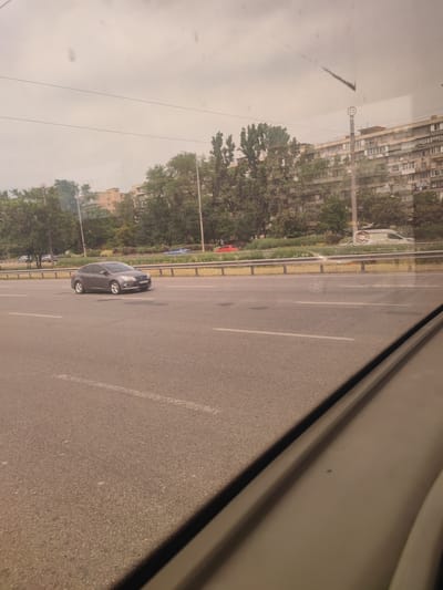 The image is an exterior, medium-shot photograph taken from a vehicle, capturing a view of a multi-lane road in Petropavlivska Borshchahivka, Ukraine. The foreground is dominated by the asphalt road, with white lane markings visible. In the mid-ground, a grey sedan is driving in the right lane, its headlights on. To the left of the road, a metal guardrail separates it from a green grassy area and a line of trees. Beyond the trees, apartment buildings are visible, indicating an urban or suburban setting. The sky is overcast with a hazy, light grey appearance. The overall scene depicts a typical daytime urban environment with ongoing traffic.