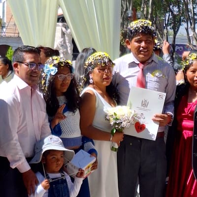 The image depicts a wedding celebration, likely in Cerro Colorado, Peru. A bride and groom are prominently featured, showered with confetti. The groom, wearing a light-colored shirt and dark pants, holds a certificate with a red heart labeled "14" attached to a bouquet of white flowers. The bride wears a white dress. Surrounding them are other people, including children, potentially family members. One child is wearing a light blue sun hat. The environment seems to be outdoors, possibly a venue with a draped or tent-like structure. Based on the image's orientation, the scene seems to have been taken on a street.