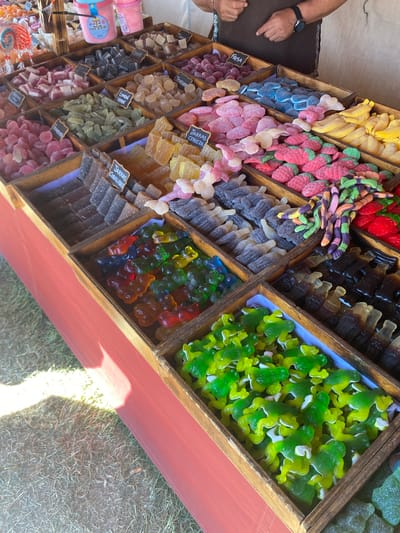 The image shows a street market stall in Llanera, Spain, featuring a wide assortment of colorful gummy candies displayed in wooden crates. The candies include various shapes and colors, such as gummy bears, worms, frogs, strawberries, and banana slices, with some labeled in Spanish. A person's hands are visible in the upper right corner, suggesting they are operating the stall. The stall is set up outdoors, with a red table covering visible in the foreground and grassy ground beneath. The lighting suggests it is daytime, possibly sunny with some shadows.