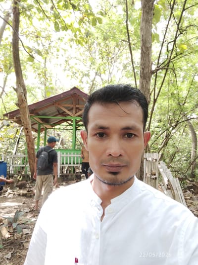 A man in a white shirt takes a selfie in a wooded area near Tu, Indonesia. In the background, a green-painted gazebo stands on a slightly elevated platform. A man wearing a backpack walks away from the camera near the gazebo. Other people are faintly visible near the gazebo. The ground is covered in dirt, rocks, and vegetation. Trees surround the scene. The date "22/05/2026" is visible in the bottom right corner of the image.
