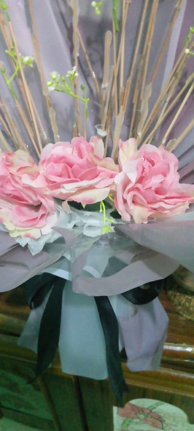 The image shows a close-up view of a bouquet of artificial flowers. The bouquet features several large pink roses with pale yellow centers, along with smaller white flowers on thin green stems. The flowers are arranged with thin, light brown reeds that have some clear, viscous material dripping down them. The bouquet is wrapped in layers of translucent gray and white material, with a black ribbon tied around the base of the wrapping. The bouquet is placed in what appears to be a light blue vase, which is partially visible at the bottom. The background is softly blurred, with hints of a wooden surface and a textured, possibly fabric, material. There are no people or animals visible in the image. The location is not determinable from the image.