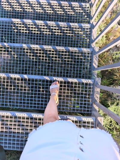 The image captures a first-person perspective of someone walking down a metal grate staircase, likely an outdoor structure. The staircase is made of gray metal with a grid pattern. To the right of the staircase, there is a metal railing and a natural environment with dry grass and some green foliage. A person's leg and foot, wearing a white sandal, are visible on the third step from the bottom. The person is also wearing a white garment, possibly a dress or skirt, with a fringed hem. The sunlight casts shadows on the staircase, indicating it is daytime. The scene does not provide specific details about the city of Shikhan, Russia, other than the potential location.