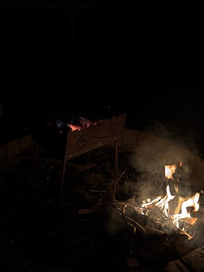 In the city of Kierwik, Poland, an outdoor fire pit is illuminated by a campfire. The fire pit is made of concrete blocks and contains burning logs and wood. Smoke billows from the fire, obscuring the surrounding area. A metal grill sits above the fire, its decorative swirl pattern visible. The scene is dark, with the fire's glow providing the primary light source. The ground around the fire pit is covered in dirt, leaves, and scattered debris.