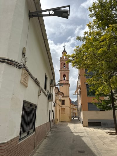 The image depicts a narrow street in Benirredrà, Spain, with a bell tower of a church visible in the distance. The street is paved with light-colored square tiles, and sunlight casts shadows from the buildings and a tree on the right side of the frame. On the left, a white building features a street sign that reads "CARRER DEL BON AIRE". There are electrical wires and conduits running along the exterior of this building, and air conditioning units are mounted on the wall. A window with black bars is visible on the lower left. The building on the right is a mix of orange and beige, with horizontal grey blinds on its windows. A green tree with light green leaves extends into the upper right portion of the image. The sky is partly cloudy. In the background, the bell tower is pink and yellow, with arched openings containing bells. Several other buildings of varying heights and colors line the street, receding into the distance. No people are present in the scene.