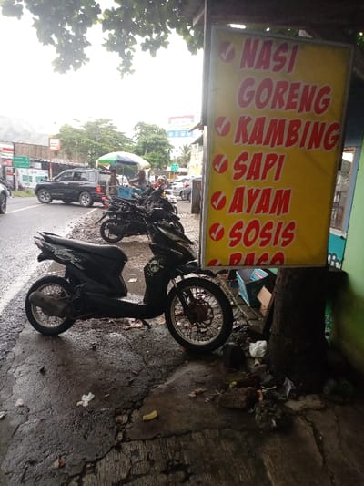 The image shows a street scene in Kecamatan Caringin, Indonesia. It is a narrow street with a line of parked motorcycles and a black SUV. On the sidewalk is a tree with a sign that reads "Nasi Goreng, Kambing, Sapi, Ayam, Sosis,  [unreadable]." There is trash and debris on the ground around the base of the tree. The sidewalk is cracked and wet. Two people are standing by the parked motorcycles, one holding an umbrella.  There is a building in the background, with a sign that is partially obscured.
