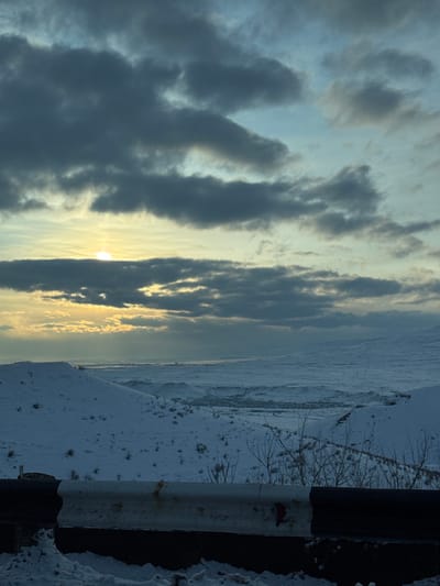 The image displays a panoramic winter landscape identified as the area surrounding Jraber, Armenia. In the immediate foreground, a white and black highway guardrail, partially encrusted with snow and ice, runs horizontally across the bottom of the frame. Behind it, sparse, dormant vegetation with bare branches protrudes from the snow. The midground features undulating hills entirely covered by a layer of pristine snow, with scattered dark patches indicating low-lying vegetation. In the distant midground, a discernible cluster of buildings, forming a small settlement, is situated within a wide, snow-covered valley. The background consists of further snow-draped hills and mountains merging with the horizon. The sky is predominantly overcast with heavy, dark grey clouds, but the sun is visible on the left side of the frame, partially obscured by clouds. It casts a localized bright yellow-orange light through an opening in the cloud cover, with faint sunrays extending downwards. No human activity or animate subjects are observed. The scene is static, under natural light, and depicts a cold environment.