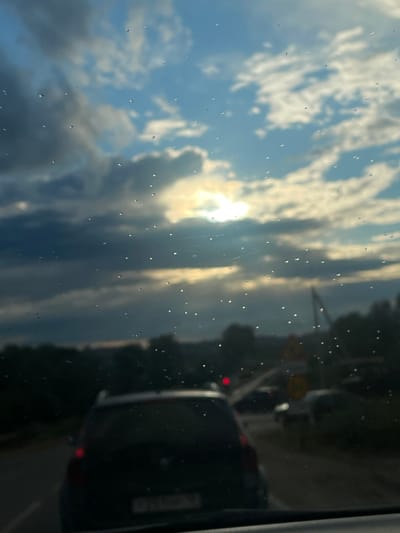 The image depicts a view through a wet window, likely from inside a vehicle. The subjects are a cloudy sky with the sun partially obscured by clouds, and a reflection of traffic on a road. We see the back of a black SUV and other vehicles in the distance. The scene is set on a road, indicated by the traffic and a yellow traffic sign visible on the right. There are numerous water droplets on the window, suggesting it is raining or has recently rained. The lighting indicates it is daytime, likely late afternoon or early evening due to the warm, diffused sunlight. The overall mood is somewhat somber due to the weather, but the sun's rays offer a hint of brightness. No discernible text is visible. The location is identified as Gavrilovka, Russia.