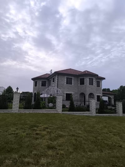 A two-story stone-clad house with a red tile roof stands behind a black metal fence with stone pillars. The house has multiple arched windows and a covered entryway with an attached garage. A well-maintained lawn is in the foreground. The sky is overcast with scattered clouds.