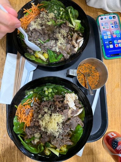 Here is a detailed description of the image:
**Scene:** A top-down shot capturing a meal setting, possibly in a restaurant or a casual dining environment.
**Subjects and Objects:**
* Two bowls of Asian-style noodles, with beef, spinach, shredded carrots, and broccoli as toppings. One bowl is being stirred with a white plastic spoon by a human hand in the lower-left corner.
* A stainless steel container filled with spices and a spoon.
* A black serving tray holding the bowls and container.
* Wooden chopsticks.
* A white napkin
* A can of Coca-Cola featuring FIFA branding.
* An iPhone with a green case displaying app icons.
**Environment:**
* The scene is set on a wooden table with a light-colored surface.
**Action:**
* Someone is in the process of eating or preparing to eat the noodle dish.
Based on the FIFA branding on the Coca-Cola can, this image might be taken during a period when FIFA events were being promoted. The overall composition suggests a casual dining experience.