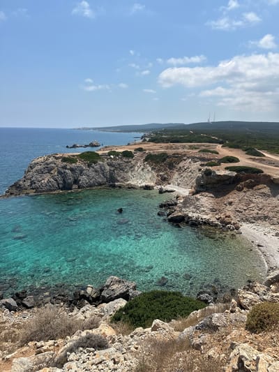 The image depicts a coastal scene in Rizokarpaso, Northern Cyprus. A rugged, rocky coastline meets clear, turquoise waters. The sea is calm with visible rocks beneath the surface. The land is characterized by arid terrain with sparse vegetation, including dry bushes and some patches of green shrubs. In the distance, hills covered in dense green foliage are visible, with a dirt road winding through them. Two tall radio towers stand prominently on the highest point of the distant hills. The sky is partly cloudy, with scattered white clouds against a pale blue backdrop. The lighting suggests a bright, sunny day. There are no people or animals visible in the frame.