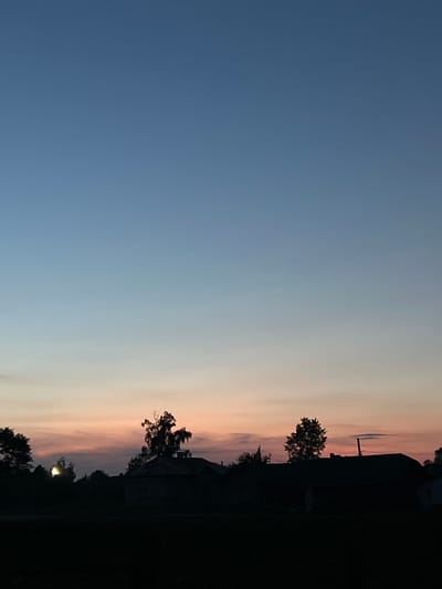 The image depicts a rural landscape at dusk, with a gradient of colors in the sky transitioning from deep blue at the top to soft orange and pink hues near the horizon. Silhouetted against the colorful sky are trees and the rooftops of buildings, suggesting a village or small settlement. The foreground is dark and indistinct, likely representing unlit fields or ground. The overall scene evokes a peaceful and serene atmosphere. The image is identified as being from Ilavche, Ukraine.